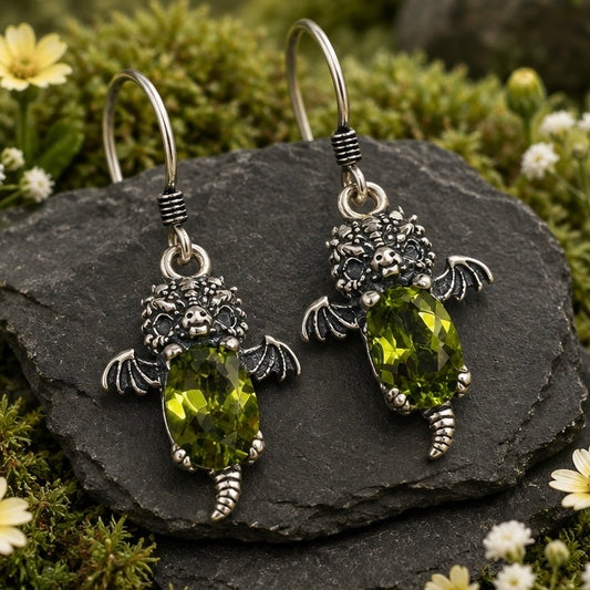 A pair of silver baby dragon drop earrings with peridot gemstone bodies, displayed on a dark slate surrounded by green moss and small white and yellow flowers.