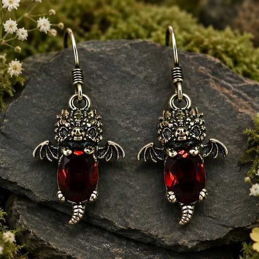 A pair of gothic-style baby dragon dangle earrings featuring silver-toned metal with black oxidized details, small  wings, and a faceted dark red gemstone, displayed on a dark stone surrounded by green moss and small white flowers.