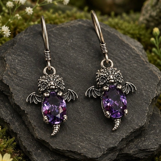 A pair of gothic-style silver baby dragon drop earrings with amethyst gemstone centers, displayed on a dark slate rock with green moss and small white flowers in the background.