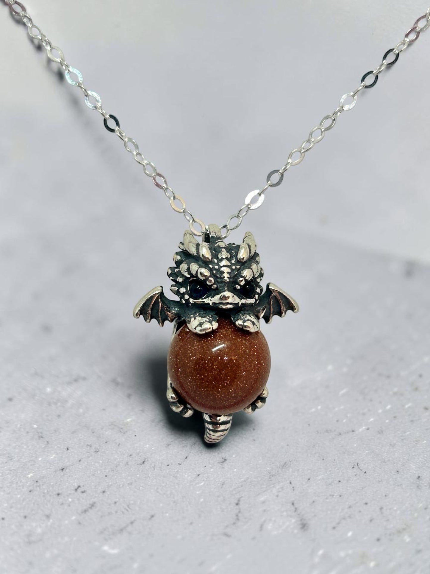 Celtic Dragon Jewellery Sterling Silver baby dragon pendant with blue eyes holding a sparkling red sandstone sphere.